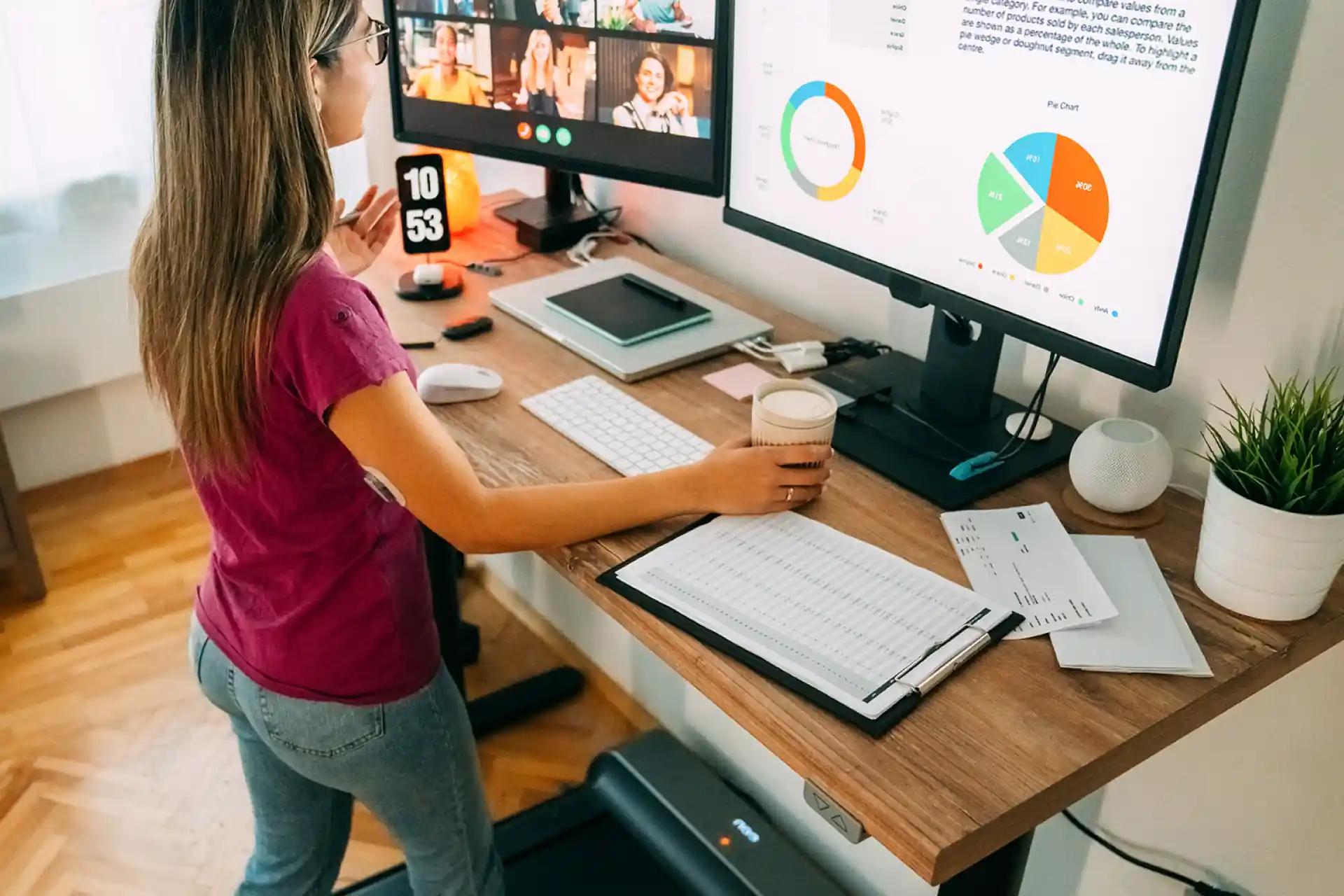 Frau an einem Laufbandtisch die an einer Videokonferenz teilnimmt während sie Daten auf einem zweiten Monitor überprüft