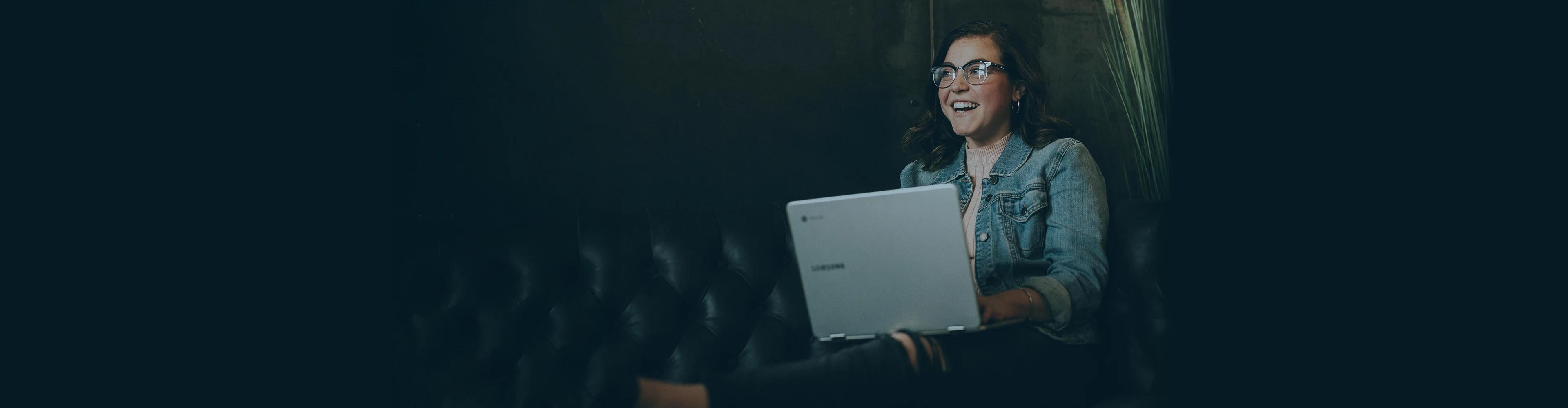 Berufstätiger mit Brille der lächelnd einen Laptop benutzt in einer modernen dunkel gestalteten Umgebung sitzt