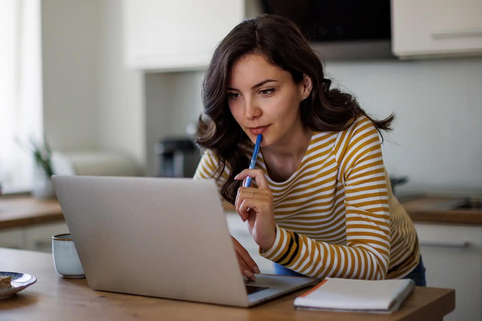 Junge Frau die von zu Hause aus mit einem Stift an den Lippen nachdenklich an einem Laptop arbeitet